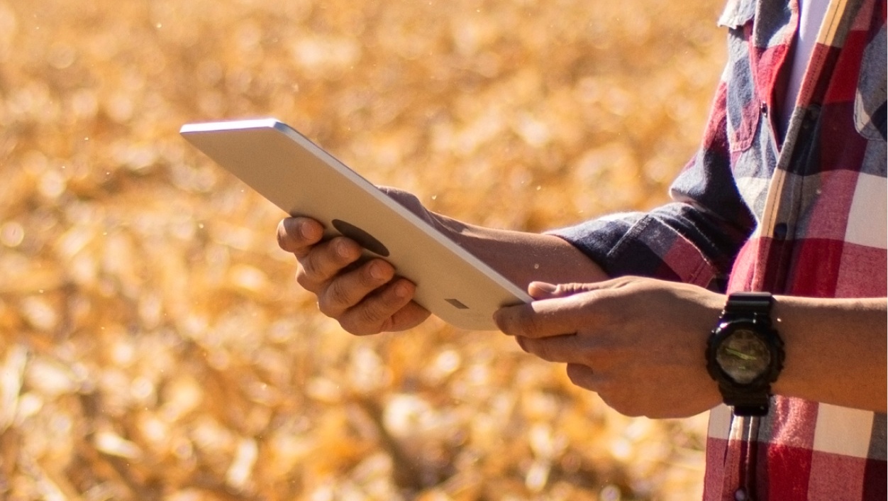 Um close lateral das mãos de uma pessoa segurando um tablet prateado. Ela veste uma camisa xadrez vermelha e azul e um relógio preto. O fundo está desfocado, mostrando um campo seco e ensolarado.